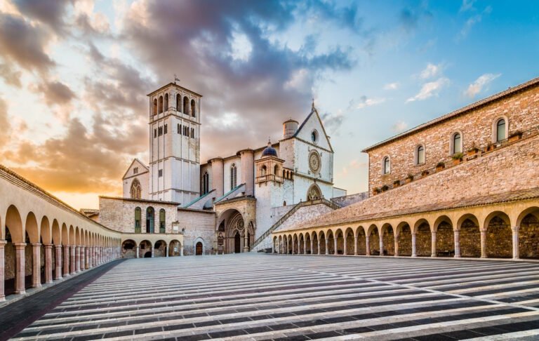 La Basilica di San Francesco ad Assisi vista dalla Piazza Inferiore deserta, con i due loggiati che convergono, alla luce del tramonto