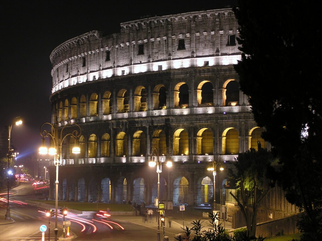 Il Colosseo a Roma di notte, con le arcate illuminate e le scie delle auto sulla strada accanto