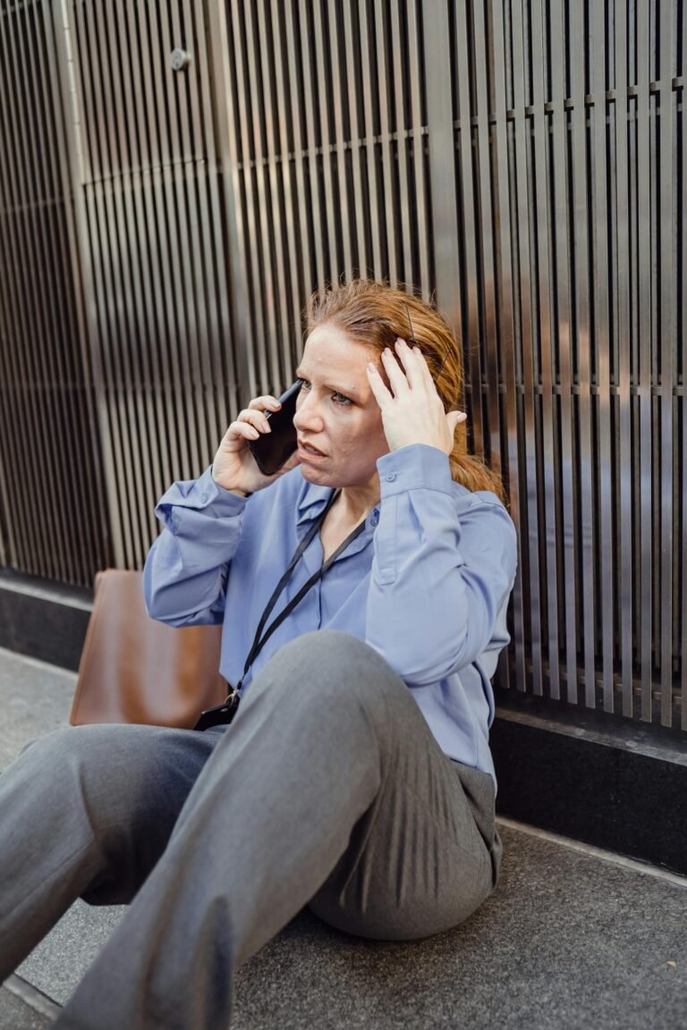 Una donna seduta sul marciapiede in una strada di città, telefono in mano, con espressione preoccupata