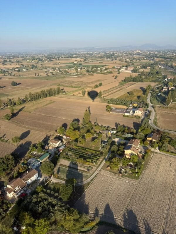 Veduta aerea della Valle Umbra al mattino, con l'ombra della mongolfiera sui campi e Assisi in lontananza